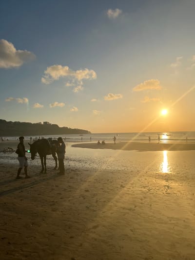 This is a photograph taken on a beach at sunset. The sun is low in the sky, casting a warm, golden light across the scene. The sky is a gradient of blue and orange, with a few scattered clouds. The beach is wet and reflective, with many footprints in the sand.
In the foreground, two people are standing next to a dark-colored animal, possibly a horse. The person in front appears to be leading the animal, while the person behind them is standing close by. Further down the beach, other people can be seen walking and enjoying the sunset. The ocean is visible in the background, with gentle waves rolling onto the shore. There is a tree-covered hillside in the distance.
The overall mood of the photograph is peaceful and serene, with a sense of relaxation and enjoyment. The time of day is clearly sunset, and the weather appears to be clear and pleasant. The presence of people and the horse suggests that this might be a tourist location where horse rides are offered. The location is Kuta Selatan, Indonesia, known for its beautiful beaches and sunsets.