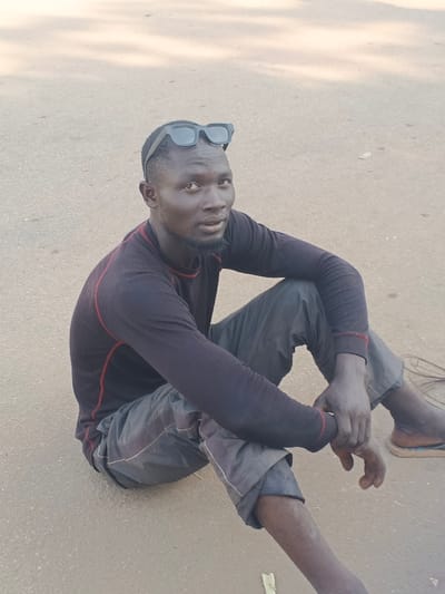 A male individual is seated directly on a light brown, possibly paved, outdoor surface in Doma, Nigeria. He has dark skin, short dark hair, and a short beard. Black, rectangular-framed sunglasses are positioned on the top of his head. He is dressed in a long-sleeved dark shirt, likely black or dark navy, featuring red stitching along the collar, shoulders, and cuffs, and dark grey trousers. His legs are bent with his knees slightly raised, and his hands are clasped or resting near his left knee. He looks directly forward with a neutral expression. His visible right foot is bare, while a light-colored sandal or flip-flop is partially visible beneath his left foot. The background consists solely of the undifferentiated light brown ground surface.