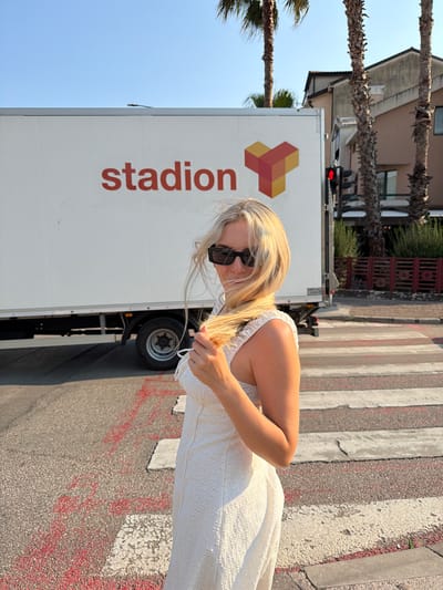 A young blonde woman wearing a white dress and sunglasses stands on a pedestrian crossing in Budva, Montenegro. She is holding a strand of her hair. Behind her is a white truck with the logo "stadion" and an abstract logo of interlocking cubes. Palm trees and buildings are visible in the background. The sun is shining, casting shadows.