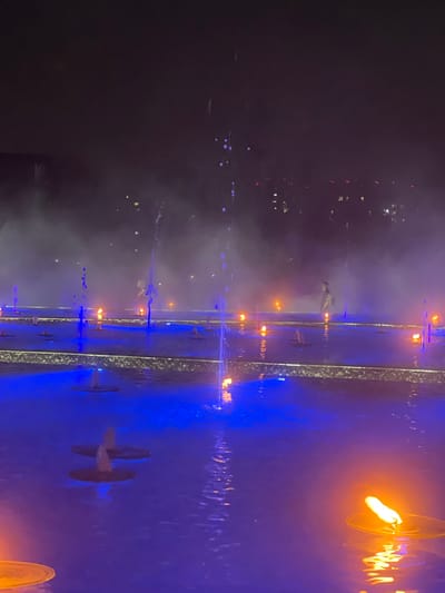 The image depicts an outdoor fountain display at night, likely in the city of Krasnodar, Russia, given the hint. The fountain features multiple water jets, some with blue lighting, creating a misty and ethereal atmosphere. Several small, circular platforms are submerged in the water, with water jets emerging from them. In the background, a hazy city skyline is visible, with lights from buildings suggesting an urban setting. There is a single person standing in the middle ground, silhouetted against the mist and lights. The foreground shows flames of what appear to be floating candles, their orange glow reflecting on the water's surface, which is predominantly illuminated by blue light from the fountain's lighting system. The overall scene is one of a modern, artistic water and light installation.