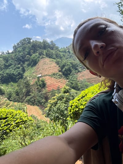 The photograph shows a person taking a selfie. The background is a landscape featuring terraced fields, common in mountainous agricultural areas. Green vegetation covers the slopes, indicating a lush environment. Sky with scattered clouds visible. Location appears to be in the Hoàng Su Phì Commune, Vietnam.