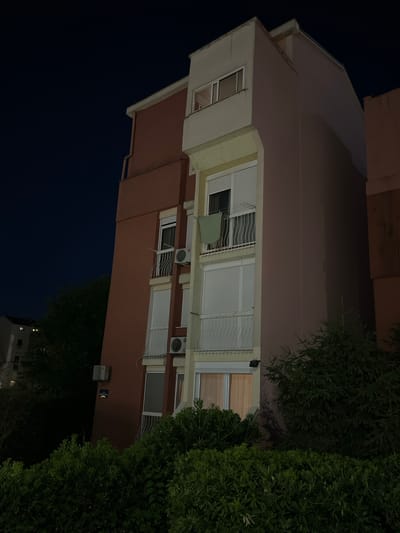The image shows the side of a multi-story apartment building at night in Budva, Montenegro. The building has a reddish-brown facade and cream-colored trim around the windows and balconies. There are several windows with white blinds, and some balconies with white railings. An air conditioning unit is visible on one of the balconies. A green cloth is hanging on a clothesline on one of the balconies. There are trees and bushes in the foreground and to the sides of the building, and another building with lit windows is visible in the distance to the left. The sky is dark blue, indicating it is nighttime. A street sign is mounted on the reddish-brown facade of the building.