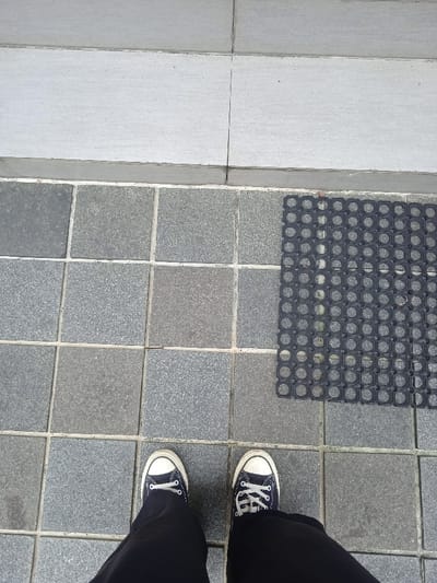The image shows a person's feet standing on a tiled floor in front of a black rubber mat. The person is wearing black pants and black and white sneakers. The tiles are a light grey color and are arranged in a grid pattern. The rubber mat is in the upper right corner of the image. The location of the image is in Surakarta, Indonesia, at coordinates -7.569 latitude and 110.811 longitude. The photo is taken from a bird's eye perspective, and the person is standing in the middle of the frame. The image is well-lit and the colors are clear.