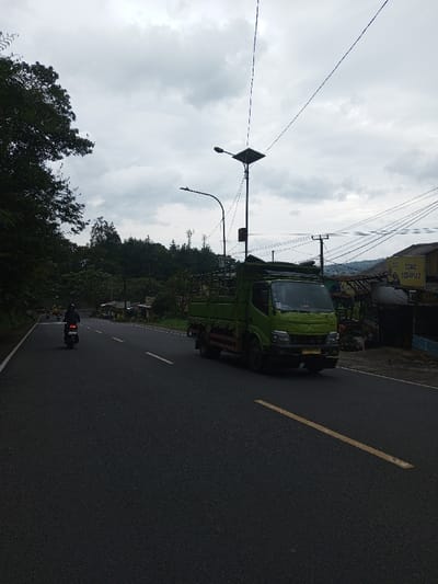 The scene depicts a two-lane asphalt road in Ciherang, Indonesia, under an overcast sky. A light green, medium-duty flatbed truck with a high-sided cargo cage is positioned in the right lane, moving towards the viewer's right. Its front bumper displays a yellow license plate. Further down the road, a solitary motorcyclist, wearing dark attire and a helmet, is operating a motorcycle in the left lane, moving away from the viewer. The road surface features a broken yellow line separating traffic directions and a solid white line marking the right shoulder. To the right of the road, a semi-permanent structure identified by a faded yellow sign reading "TOKO SEDAYU 2" is visible. Multiple utility poles with a dense network of overhead electrical and communication wires are present near the structure. One prominent streetlight post features a solar panel array atop its fixture. The left side of the road is bordered by dense green foliage, transitioning into tree-covered hills in the distance, where telecommunication towers are discernible. The sky is uniformly grey and heavily clouded.