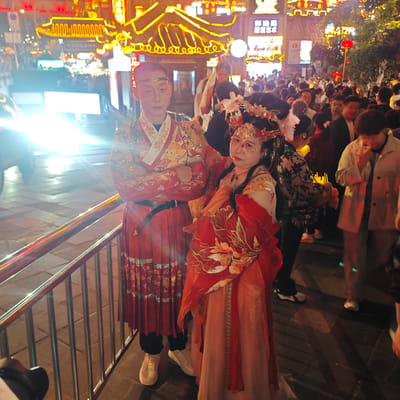 In Yuzhong District, China, a man and woman dressed in traditional Chinese clothing pose for a photo near a metal railing. The man wears a red and gold patterned robe with a black belt, black pants, and white sneakers. He has a silver headband and his arms are crossed. The woman wears a long, flowing red and peach dress with floral embroidery, as well as a golden headpiece and sparkling facial decorations. A crowd of people is gathered behind them in the background. Streetlights and neon signs illuminate the scene.