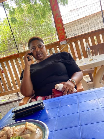 The image shows a woman sitting at a table outdoors in Dar es Salaam, Tanzania, at the coordinates -6.77 latitude and 39.225 longitude. The woman is wearing a black shirt and holding a cell phone to her ear. Her left hand is resting on the table, and she is wearing bracelets on her wrist. She is also wearing a red purse and a black wallet. The table is blue and has a plate of food on it. There is a wooden fence behind the woman, and a green tree can be seen through the fence. The woman appears to be on the phone and looking at the camera. The scene appears to be during the day. The image was likely taken in a local eatery, as the surrounding area suggests a casual, outdoor setting.