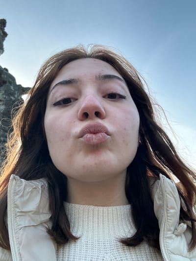 A young woman with dark brown hair is centered in a low-angle selfie. Her lips are pursed. She has fair skin. She is wearing a cream-colored sweater and a puffy cream-colored jacket. The sky is light blue and visible in the background, along with a glimpse of rocky terrain. The image is bright and slightly tilted.