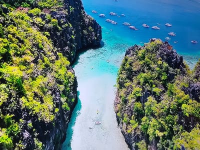 This is an aerial drone shot of a tropical landscape. Two large, jungle-covered rock formations flank a narrow, turquoise channel of water. Several small boats, likely for tourism, are anchored in the deeper, blue ocean beyond the channel. The water closer to the shore is so clear that the sandy bottom is visible. In the channel, two people are kayaking, and another small boat is visible. The sun is shining brightly, casting shadows on the rocks and illuminating the water. The location is not in Sofia, Bulgaria, as it is a tropical island setting.