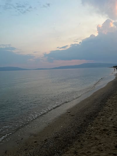 This image depicts a beach scene in Kariani, Greece, during twilight. The sky transitions from a pale blue near the horizon to a soft lavender and pink at the top, with scattered clouds. A sliver of the moon is visible in the upper left quadrant. The sea is calm, with gentle waves lapping the shore. The beach itself is a mix of sand and pebbles, with some faint footprints visible on the sand. In the distance, a silhouetted coastline with mountains can be seen. On the right side of the image, along the waterline, two small figures of people are visible walking.