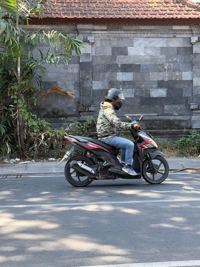 A person wearing a camouflage jacket, blue jeans, and a black helmet is riding a black and red scooter on a paved road. The scooter has a visible exhaust pipe and a rear fender. The rider's left foot is on the footboard, and their right foot is visible touching the ground near the rear wheel. The scooter is moving towards the right of the frame. In the background, a grey stone wall with decorative elements and an orange tiled roof can be seen. Lush green vegetation, including palm fronds, grows along the base of the wall. The road surface is asphalt with white lane markings and is partially covered in dappled sunlight and shadows. The scene appears to be a street in North Kuta, Indonesia, given the architectural style of the wall and roof, and the presence of tropical vegetation.