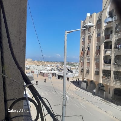 The image, captured from an elevated viewpoint in Khan Yunis, Palestinian Territory, presents a daytime scene under a clear blue sky. In the foreground, dark cables cross the frame, partially obscuring a light-colored building edge on the left. A light gray street light pole stands prominently in the mid-ground, casting a shadow to its left.
Below the street light, a paved road with a pedestrian crosswalk extends horizontally. Individuals are visible on and adjacent to the road; one person walks left-to-right on the crosswalk, another stands near a building to the far right, and a small group moves on the unpaved ground to the right of the road.
To the right of the street light and road, a multi-story residential building with a light-beige facade, balconies, and patterned window surrounds occupies the right side of the frame. Laundry or fabrics hang from multiple windows and balconies, and a white water tank is visible on its roof.
To the left of the street and extending into the background, an expansive area of makeshift shelters, tents, and rudimentary structures is spread across sandy, uneven terrain, indicative of an informal settlement or refugee camp. Several people are observed moving within this area. Beyond the camp, large mounds of earth or sand are present, with the distant Mediterranean Sea visible on the horizon. The scene depicts a populated area blending urban residential structures with an extensive, temporary settlement.