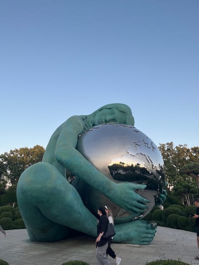 A large, teal-colored sculpture of a kneeling figure cradling a mirrored globe is the central focus of the image. The figure appears to be female, with its head resting on the globe. The globe reflects the surrounding environment, showing trees, people, and a clear blue sky with a faint sunset glow. In the foreground, three people are walking on a paved pathway. One person on the left is partially visible, wearing a grey hoodie. Two people in the center are walking together; one is wearing a white top and dark pants, and the other is wearing a black top and grey pants, carrying a black tote bag. A man in a black t-shirt and shorts stands to the right, looking at his phone. The sculpture is set within a manicured garden with lush green bushes and trees. The location is the Monument "The Motherland of the World" or "Mother Earth" in Krasnodar, Russia.