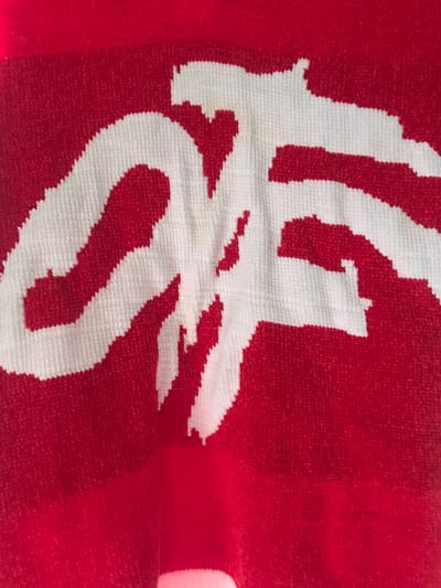 The media file displays a close-up image of a red textile, likely a towel or blanket, featuring a large, stylized white design. The fabric has a textured, looped or pile surface, characteristic of terrycloth.
**Subjects:** The primary subject is a textile item. No people or animals are visible.
**Objects:** The main object is a red and white woven fabric, which appears to be a blanket or towel.
**Scene/Setting:** The scene is a tight shot of the fabric, offering no discernible background or environmental setting beyond the textile itself.
**Activity or Event:** No activity or event is depicted; the image is a still shot of an object.
**Notable Details:**
* **Colors:** The dominant colors are vibrant red and bright white.
* **Texture:** The fabric exhibits a clear, soft, and plush texture, typical of woven materials like towels, with distinct loops. The edges of the white design are slightly jagged, reflecting the weaving process.
* **Design:** A prominent, large, white stylized script or logo is woven into the red background. This design strongly resembles the classic "Coca-Cola" script, appearing to show parts of the letters "C", "o", "c", "a", and "Cola" with its distinctive cursive flow and connected letters. The design is central and occupies a significant portion of the frame.
* **Condition:** Faint, brownish-yellow stains are visible on the white portion of the design, particularly in the central-left area. The fabric itself shows slight folds and creases.
* **Lighting:** The lighting appears even, illuminating the texture and colors clearly.
* **Location Cues:** No explicit location cues are present within the image to suggest Onitsha, Nigeria, beyond the provided context.
**Visible Text:** The white design is a highly stylized script, which is identifiable as the "Coca-Cola" logo.