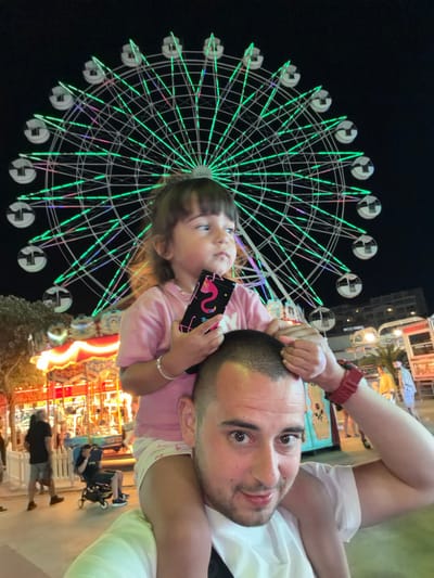 A man with short brown hair and stubble is taking a selfie at night in what appears to be an amusement park in Nesebar, Bulgaria. He is wearing a white t-shirt. A young girl with brown hair, wearing a pink t-shirt and white shorts, is sitting on his shoulders. She is holding a black phone with a pink snake design. In the background, a large Ferris wheel is illuminated with green lights. To the left of the Ferris wheel, a carousel is visible with bright, colorful lights. Several people are seen in the background, including a man in a black t-shirt and shorts, and a person in a stroller. The overall scene depicts a festive, nighttime amusement park atmosphere.
