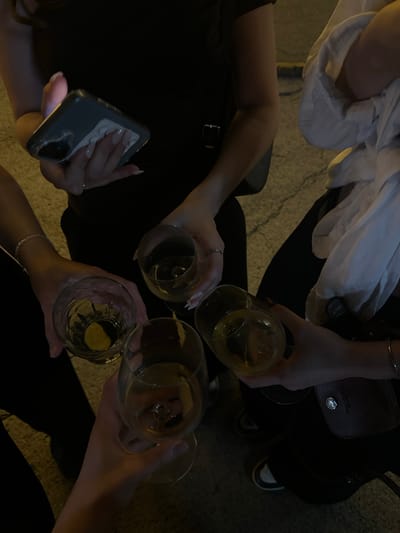 This is a close-up, top-down medium shot of four individuals gathered outdoors at night, likely on a cobblestone street given the texture of the ground. The focus is on their hands holding glasses of amber-colored liquid, suggesting they are toasting or celebrating. One person on the left is holding a smartphone, its screen illuminated. Another person in the center-right is wearing a white shirt. The individuals appear to be in Novi Sad, Serbia, though the specific location within the city is not discernible from the image alone. The overall mood is convivial and social.