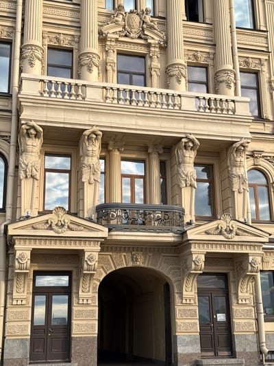 The image shows the facade of a historic building in Saint Petersburg, Russia, likely a palace or a grand apartment building. The architectural style is ornate and classical, with a prominent beige stone facade. Key features include large Corinthian columns, intricate carvings, decorative reliefs, and a balcony with a stone balustrade. There are two large statues of Atlantes holding up the upper structure, located on either side of a central balcony. The windows are tall and rectangular, with some reflecting the sky and clouds. Below the balcony, there is an arched entryway leading into the building. The entrance is flanked by decorative pilasters and topped with a pediment. The overall impression is one of grandeur and historical significance, typical of Saint Petersburg's imperial architecture.