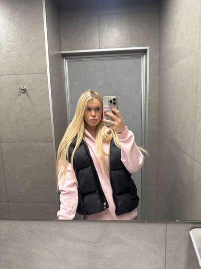The media file shows a mirror selfie taken by a young woman with long blonde hair. She is wearing a pink hoodie and a black puffer vest. Her nails are painted red. She is holding a smartphone in her right hand, angled to capture her reflection. The background appears to be a bathroom with grey tiled walls and a glass shower door. The lighting suggests an indoor setting, possibly during the daytime. There is no visible text in the image. The setting and the woman's attire do not provide specific cues about the time of day or weather. The woman's expression is neutral, and she is looking directly at the camera. The location context of Saint Petersburg, Russia, is provided externally and is not discernible from the image itself.