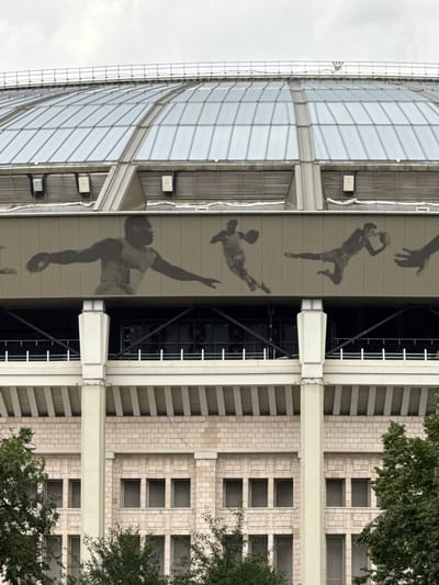The image depicts the exterior of the Luzhniki Stadium in Moscow, Russia. The stadium features a distinctive domed roof constructed of glass panels. Below the roof, a beige-colored band runs along the exterior, adorned with large, dark silhouette images of athletes engaged in various sports, including what appears to be discus throwing and basketball. The stadium's facade is characterized by large, pale stone blocks and rows of windows. Trees and foliage are visible in the foreground, suggesting an outdoor setting. The sky is overcast.