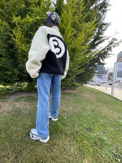 The image shows a person standing on a grassy area with a tree to their left. The person is wearing a white and black jacket with a large "8" ball design on the back, blue jeans, and white sneakers with black detailing. They are also wearing a black and white bandana. The location appears to be Ushuaia, Argentina, judging by the presence of signage visible in the background. The person is facing away from the camera and towards the signage and the street in the distance. The scene appears to be outdoors on a partly sunny day.