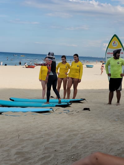 A group of people are on a sandy beach in Đà Nẵng, Vietnam, with the ocean in the background. Three women in yellow shirts stand on the beach, and one woman in a black and red rash guard gestures towards them. A man in a neon green shirt and a hat stands to the right, appearing to be a coach. Two blue surfboards are laid out on the sand in front of the group. The sky is partly cloudy, and the ocean is calm with several boats and people in it. A yellow and green sign stands on the right side of the image with Vietnamese text.