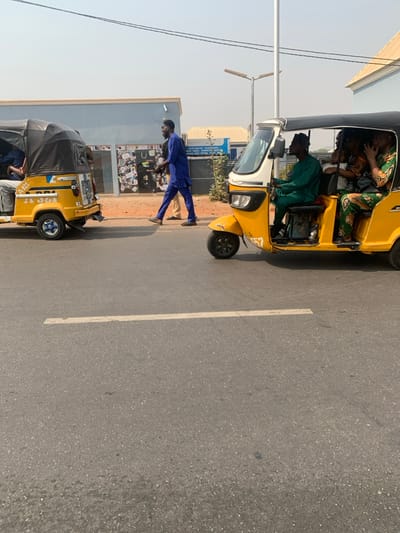 This image captures a street scene in Adankolo, Nigeria, under a clear, bright sky. The foreground features an asphalt road with a faded white painted line. Two yellow motorized tricycles, known as Keke Napeps, occupy the road.
On the right, a Keke Napep is in motion, moving from right to left. Its male driver, wearing a dark green traditional outfit, is visible at the controls. The passenger compartment contains at least three individuals; a female passenger in a green, orange, and black patterned traditional dress is prominent, with her right hand raised near her face.
On the left, another yellow Keke Napep is partially visible from the rear, either stationary or moving slowly away from the viewer. Its rear plate area displays "A-321". At least two passengers are discernible within its canopy.
Between the two vehicles, on a dirt shoulder adjacent to the road, a male pedestrian walks from right to left. He is dressed in a royal blue traditional Nigerian garment. The lower legs of another person are partially visible behind him.
In the background, a light blue building with a black roof and multiple windowed storefronts is visible on the left. Further behind and to the right, a second, lighter blue building with a cream roof is present. A tall, light-colored street lamp pole with two horizontal fixtures stands near the center-right, extending upwards. Multiple dark overhead utility lines span across the upper portion of the frame. The distant sky appears light blue and slightly hazy.