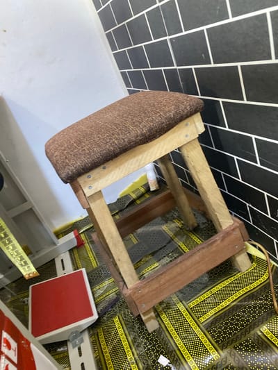 This media file shows a close-up view of a small, homemade stool with a brown upholstered seat, constructed from light-colored wooden planks. The stool's frame is visible, with screws securing the wooden pieces. Behind the stool, a black and white tiled wall forms a backdrop, with the tiles laid in a staggered pattern.
Below the stool, a patterned fabric in yellow and black is laid out, featuring a repeating geometric design. To the right of the stool, a white metal bracket with red accents is attached to the wall, possibly part of a shelving unit. A yellow measuring tape with black markings is also visible in the lower right portion of the frame, draped over a surface.
The image appears to be taken indoors, with the lighting suggesting daytime. The overall impression is that of a workshop or domestic space where crafts or repairs might be taking place. The location context provided is Jalingo, Nigeria. No people are visible in the frame. There is no discernible text within the image.