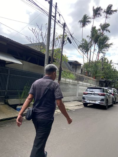 The image shows a man walking on a paved road in Bandung, Indonesia, at the coordinates -6.939 latitude, 107.572 longitude. He is wearing a black t-shirt with "Chanel Paris" printed on it, black pants, and a black shoulder bag. He is walking away from the camera and towards the right side of the frame. There is a silver car parked on the right side of the road, facing the camera. The car is parked behind the man and is partially obscured by him. The background shows several palm trees and a residential building with a fence. The scene is overcast, and there are power lines running overhead.