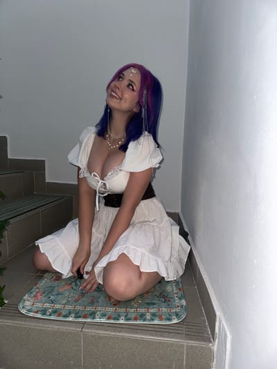 A female kneels on a patterned rug placed on a tile floor. She is dressed in a white dress with lace trim and a dark belt. She has blue and purple hair, and a jeweled headpiece. The woman is smiling and looking to the left. The background includes a white wall, steps, and a plant. The location is implied to be City of Zagreb, Croatia.