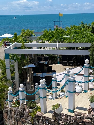 The image depicts an outdoor seating area overlooking a body of water, likely the Black Sea given the context of Sochi, Russia. The foreground features a white railing with turquoise accents and chains, leading down a set of steps adorned with potted plants. Beyond this, a covered dining area with dark wicker chairs and tables is visible, with a patio heater standing prominently. In the midground, a white pergola frames the view towards the sea. The background shows a beach with a few sunbathers and umbrellas, followed by the expansive blue water. A yellow flag flies atop a lifeguard station on the water's edge, and a small boat can be seen in the distance. The sky is a clear blue with scattered white clouds. The overall scene suggests a resort or restaurant setting, possibly in the Adler district of Sochi, known for its coastal attractions.