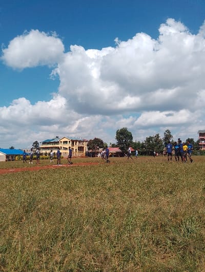 The image depicts a grassy field with several people in the distance, playing soccer. There are several buildings in the background, including a two-story building with a yellow facade, and a single-story building with a blue roof. The field is covered in dry, brown grass with a small patch of reddish-brown dirt in the foreground. The sky is clear and blue with several large, fluffy white clouds. The image was taken in Rungiri, Kenya, at coordinates -1.249 latitude and 36.692 longitude.