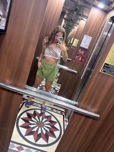An individual is taking a mirror selfie inside an elevator. The individual is a woman with curly brown hair, wearing a light pink crop top and green shorts. She is holding a smartphone in her right hand, capturing her reflection. The elevator interior features wood-paneled walls, a polished metal handrail, and a decorative floor tile with a starburst pattern. Posters are visible on the elevator walls, including one with text in Vietnamese that reads "NỮO OHN 000,002" and another with "HOTLINE 24/7: 1900". The presence of Vietnamese text and currency notation suggests the location is in Vietnam. Considering the context provided by the text on the posters and the typical branding found in hotels in Hanoi, it is highly probable that this elevator is located in a hotel in Hanoi, Vietnam.