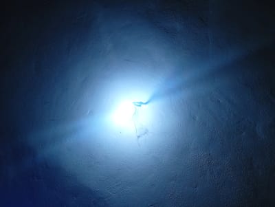 A bright, blue light emanates from the center of a dark, textured surface. The light is intensely focused, creating a halo effect and casting faint shadows and highlights on the textured background. The surface appears to be a wall or ceiling with a rough, uneven finish. No people or other objects are visible in the image. The image appears to be taken in a dimly lit environment, with the blue light being the primary source of illumination.