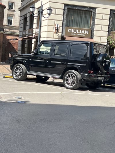 The image shows a black Mercedes-Benz G-Class parked on a street in Sofia, Bulgaria. The car is positioned at an angle, with its rear to the right of the frame. The vehicle has a spare tire mounted on the back, and its license plate reads "CB 9919 XO".

Behind the car, a building with beige stone facade is visible. A sign on the building reads "GIULIAN LUXURY COLLECTION". The street is paved with asphalt and there are some manhole covers visible on the ground. The scene appears to be in a commercial or urban area, with a lamppost also present. The time of day appears to be daytime, with the sun casting shadows. No people are visible in the image.