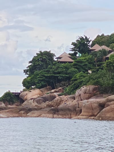 This media file shows a serene coastal scene in Baan Chaweng Noi, Thailand. The foreground is dominated by the calm, rippling surface of the ocean, reflecting the overcast sky. Beyond the water, a rocky shoreline transitions into a lush green hillside. Large, weathered boulders, varying in shades of brown and grey, form the natural embankment.
Perched atop the hillside are several structures with distinctive brown, conical roofs, suggesting resort accommodations or private villas. These buildings are nestled amongst dense, vibrant green trees, creating a harmonious blend of architecture and nature. A wooden walkway or bridge is visible on the left side, leading towards the structures.
The sky is filled with soft, diffused clouds, indicating either early morning or late afternoon, or possibly a generally cloudy day. The weather appears mild, with no harsh sunlight. A lone snorkeler, with a snorkel and mask visible, is in the water, enjoying the ocean. The overall atmosphere is peaceful and tranquil, evoking a sense of relaxation and escape. There is no visible text in the media file.