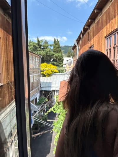 A person's dark hair, obscuring their face, dominates the foreground of the image, which appears to be taken from inside a building looking out. The person's back is turned towards the camera, with their hair falling over their shoulders.

The view outside reveals a narrow courtyard between two buildings. The building on the right is a wooden structure with a weathered, orange-brown facade and wooden-framed windows. The building on the left, partially visible, also appears to be made of wood with balconies.

Beyond these buildings, a bright yellow flowering bush or tree is visible, contrasting with the green foliage of other trees. The sky is clear and blue with some wispy white clouds. The scene suggests an urban environment, likely in Tbilisi, Georgia, given the distinctive architecture of the buildings, characterized by wooden facades and balconies, a common feature in older districts like Sololaki or Avlabari. The presence of electrical wires strung across the courtyard further indicates an urban setting. The image captures a moment of quiet observation, with the individual seemingly gazing out at the street or courtyard below.