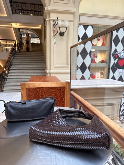 This is a media file of two handbags hanging on a metal pole in what appears to be a retail store. The larger bag is brown and woven, while the smaller bag is black and made of smooth leather. The scene is brightly lit, suggesting it is daytime. In the background, there are glimpses of a store interior, including a staircase with ornate railings and displays featuring playing card motifs. The overall impression is one of a high-end boutique. There is visible text on the black bag which appears to be a logo, possibly "MIU MIU." The image is oriented such that the bags are hanging from the top, and the rest of the store environment recedes into the distance.