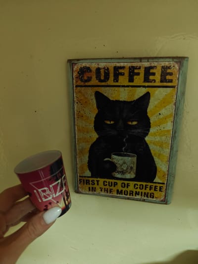 A close-up shot shows a hand with white nail polish holding a cylindrical souvenir cup with the word "IBIZA" and a sunset scene on it. To the right of the cup is a framed vintage-style metal sign depicting a black cat with yellow eyes holding a cup of coffee, with the text "COFFEE" at the top and "FIRST CUP OF COFFEE IN THE MORNING" at the bottom. The background is a plain, light-colored wall. This scene does not depict the city of Dubai, United Arab Emirates.