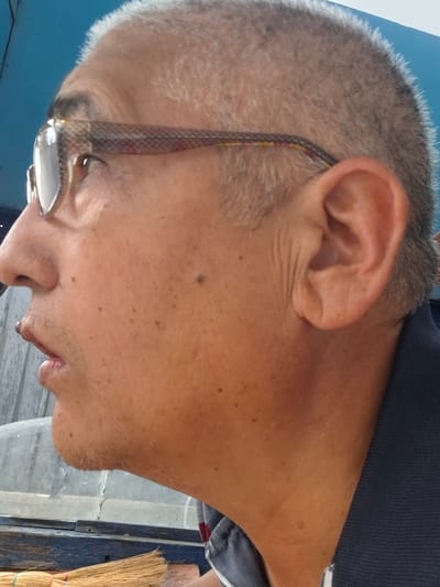 The image shows a close-up, profile view of an individual's face, from the nose to the ear and neck, with a blue background. The individual, who appears to be an adult male, has fair skin with freckles and moles. He is wearing eyeglasses with a patterned frame. His hair is short and graying. His mouth is slightly open, revealing his lips and a small portion of his teeth. A mole is visible on his left cheek, and another smaller one below his left eye. The background is a solid blue color, and a blurry element that could be a wooden surface and a broom is visible at the bottom of the frame. The scene does not provide any specific information that would allow for identification of the location within Almaty, Kazakhstan.