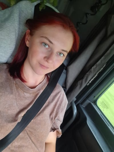 A young woman with bright red hair and light green eyes is seated in the driver's seat of a vehicle, likely a truck, given the interior details visible. She is wearing a light brown, textured t-shirt and a black seatbelt is fastened across her chest. Her hair is partially pulled back, with some strands framing her face. She is looking towards the camera with a gentle smile. The background shows the interior of the vehicle, including dark grey curtains, a black coiled cable, and part of the dashboard. A green-tinted window is visible on the right, with some text on it, possibly a manufacturer's label. The environment appears to be an urban or roadside setting, as indicated by the bright green glare from outside, suggesting daylight. The specific location in Кабачигурт, Russia, is not discernible from the visual information in the image.