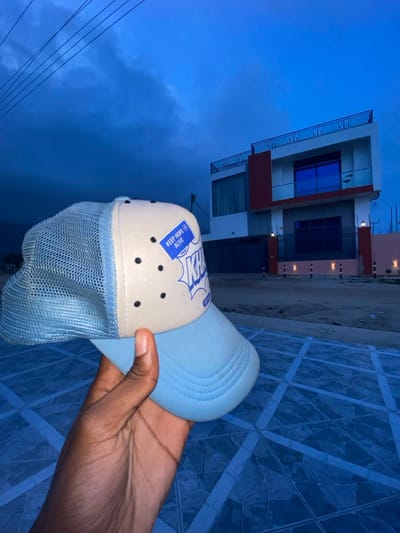 A hand holds a light blue and beige trucker hat against a backdrop of a modern two-story building. The hat has a mesh front and a blue patch with the words "Keep Hope Alive". The building features white walls with red accents and blue reflective windows. The ground has a patterned tile surface. The sky is a dark blue, suggesting either dusk or dawn. Power lines are visible against the sky on the left side of the frame.