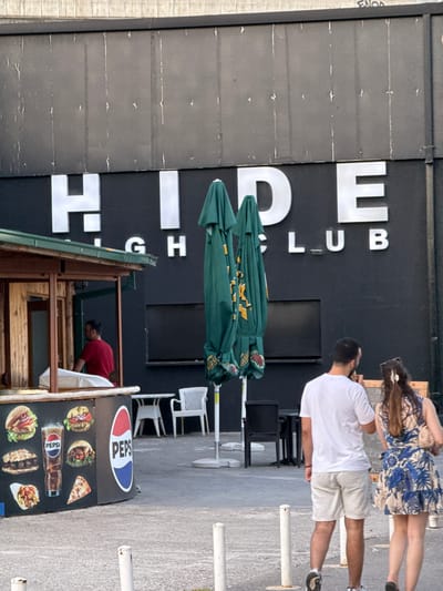 In Budva, Montenegro, a building with the illuminated sign "HIDE CLUB" is visible. In the foreground, there is a food stand with images of burgers, a glass of Pepsi, and a slice of pizza. Two people, a man and a woman, are walking away from the camera towards the right side of the frame. Several green umbrellas are stacked to the left of the building. In the background, there are white tables and chairs. The overall environment suggests an outdoor area of a venue or establishment.