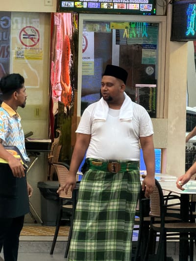 A group of people are standing in front of a shop in Kuala Lumpur, Malaysia. The shop appears to be a money exchange business, with currency displayed on a screen above the entrance. Two men are in the foreground, one on the left wearing a patterned shirt and the other on the right wearing a white t-shirt and a sarong. The man on the right has a towel draped around his neck. A woman's hand is visible in the background, holding a small white object. There are tables and chairs in front of the shop, suggesting it is an outdoor seating area. The environment appears to be urban, with buildings and signs visible in the background.