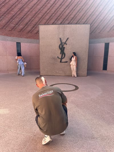 A high-angle, vertically oriented photograph captures three people in a sunlit modern architectural setting.
On the right side of the image, a man is seen from behind, bending over and looking downwards. He wears a dark olive-green t-shirt with "Balenciaga" printed in white and red across the upper back. His short hair is visible, and his right foot, wearing a white and grey sneaker, is positioned on a light grey, speckled tiled ground that dominates the right and central-right portions of the frame. Thin white lines form a subtle grid pattern on the ground, and a dark circular design is partially visible near the man.
In the center of the image, standing upright against a light-grey stone wall, is a woman with long dark hair. She is dressed in a light-colored, possibly beige or white, two-piece outfit, featuring a halter-neck top and wide-leg trousers. A large black 'YSL' logo is prominently affixed to the stone wall directly beside her.
Towards the left side of the image, further in the background, another woman is partially visible from behind. She is wearing a white top and light blue trousers or overalls and carries a bag over her shoulder. The architectural surroundings include a textured reddish-brown brick wall on the far left, and additional light-grey stone surfaces. The entire scene is brightly illuminated by natural daylight, indicating clear weather. The presence of the 'YSL' logo is a strong indicator of a location associated with Yves Saint Laurent, fitting the context of Marrakesh, Morocco.