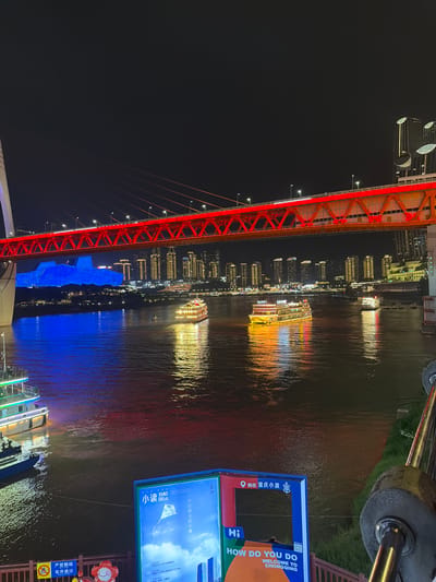 This image captures a nighttime view of the Yangtze River in Yuzhong District, Chongqing, China. A brightly lit red suspension bridge spans across the dark water, with its illuminated cables stretching upwards. Below the bridge, a large building is illuminated in blue. Several boats with golden lights are on the river, reflecting their illumination on the water's surface. In the background, a city skyline with many illuminated buildings is visible. In the foreground, there are two large signs. One sign is primarily blue with white text and a kite design. The other sign has a red frame and the text "HOW DO YOU DO WELCOME TO CHONGQING" in orange and white letters. The river appears dark and reflective, with ripples disturbing the water's surface. The sky above is black, indicating nighttime. The overall scene is a vibrant urban landscape at night, dominated by artificial lights.