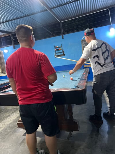 In Altagracia, Venezuela, two men are engaged in a game of billiards. The man on the left, facing away from the camera, wears a red t-shirt and black shorts. He is positioned near a billiards table, holding a cue stick. The man on the right, also facing away, wears a white t-shirt with a black logo on the back and dark pants. He is also at the billiards table, with a cue stick extended towards the balls. The billiards table is positioned under a corrugated metal roof structure. The background features a vibrant blue wall adorned with a rack of billiard balls and a small scoreboard. The lighting suggests an indoor or covered outdoor setting, possibly an entertainment venue.