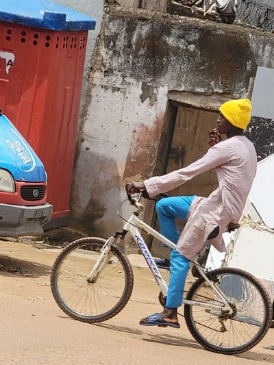 A street scene in Maiburiji, Nigeria, unfolds under bright daylight. A dark-skinned adult male, wearing a yellow beanie, a light beige or pinkish-brown long-sleeved tunic, bright blue pants, and blue open-toed sandals, is seated on a white "KED BIKE" bicycle. His right hand is near his mouth, and he looks to the right. The bicycle is stationary or moving slowly, with its front wheel visible on the unpaved ground.
To the left of the cyclist, the front portion of a light-blue vehicle, likely a van, is visible, featuring "Pure Bliss" branding on its bonnet and a red grille. Directly behind the vehicle and to the left of the cyclist, stands a tall, red, rectangular structure with a blue top, appearing to be a portable utility unit or public restroom, marked with a white "A" and multiple ventilation holes.
The background consists of a heavily weathered wall, possibly concrete or stucco, displaying significant peeling paint, discoloration, and structural wear. An open doorway is visible within this wall. The ground is an unpaved, reddish-brown dirt surface, showing minor undulations and sparse green vegetation.