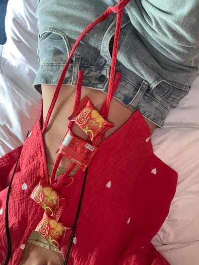 In the city of Mù Cang Chải, Vietnam, an overhead perspective captures a person lying on a white bed sheet, from the mid-torso to the upper thighs. The individual wears light-wash denim shorts and a red, textured garment adorned with small white heart embroidery. Beneath this, a light olive-green bralette and a delicate gold-toned necklace are visible. Red braided cords, either integrated into the red garment or acting as an accessory, are positioned across the upper chest. Five red snack packages, branded "Gác" with images of golden crackers and a reddish fruit, are present; four are face-up on the red garment, held by the cords, and one is reversed, showing a barcode and text. Bare skin is visible above the denim shorts' waistband. The surrounding environment is characterized by rumpled white bedding.