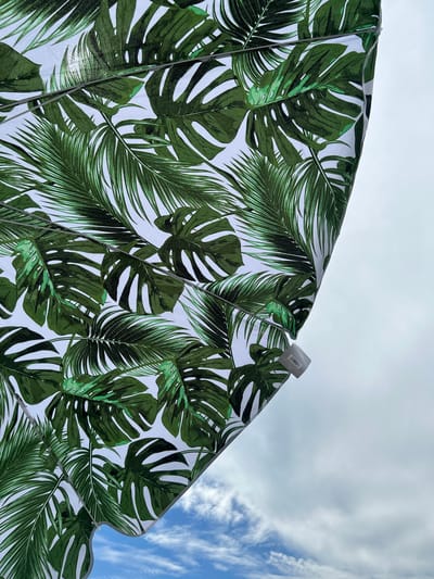 The image shows a close-up view of a tropical-patterned umbrella, featuring large green Monstera and palm leaves on a white background. The umbrella is angled upwards, with a portion of the sky visible in the upper right and lower portions of the frame. The sky is overcast with some patches of blue sky and scattered white clouds. There is a white plastic clip attached to the edge of the umbrella, and a small white tag with a barcode is also visible. The scene appears to be outdoors, possibly at a beach or park, and the overcast sky suggests a cloudy day. There are no people or animals visible in the image. The location is not specified in the image itself.