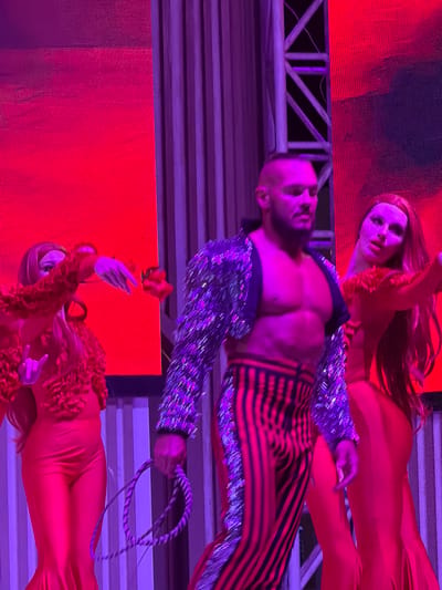 A man with a shaved head and beard, wearing a sparkly jacket and striped pants, stands on a stage. Two women in red outfits flank him, their faces obscured by shadows and lighting. The woman on the left gestures with her hands, while the woman on the right looks towards the camera. The stage features a vibrant red backdrop and purple lighting, suggesting a performance or event. The context of Sharm El Sheikh, Egypt, is not visually evident in the image itself, but the description implies a setting within that city.