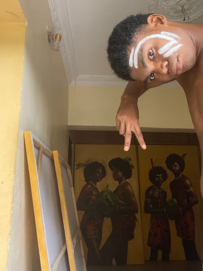 A vertically inverted photograph depicts a dark-skinned male with close-cropped, dark hair and white painted marks on his face. He is making a peace sign with his right hand. Behind him are two framed paintings of dark-skinned individuals. In the foreground are two unmounted canvases. The location appears to be inside a building with light yellow walls. The specific location within the city of Ado, Nigeria cannot be determined from the image.